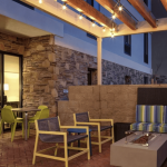 A vibrant dog park bathed in sunlight features abundant greenery with mature trees, offering ample shade for leisurely outdoor activities. In the distance is a well-equipped playground that presents an additional activity area. An inviting picnic table spreads out nearby, ideal for enjoying meals al fresco or taking relaxing breaks from play. A neatly-placed trash bin aids in maintaining the park's cleanliness for everyone's enjoyment. Dominating the scene is a generously wide dirt path gracefully curving through the expanse of grass, promising exploratory walks and lively fetch games.